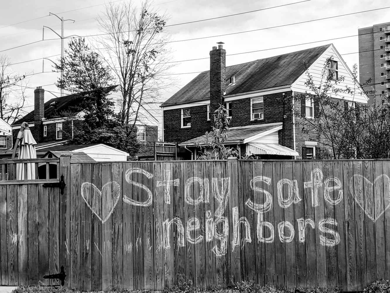 Grayscale photo of wooden fence, written on in chalk to say, "Stay safe neighbors" with two hearts surrounding the text